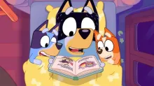 A still from the animated series Bluey showing Bluey's father reading to her and her sister.